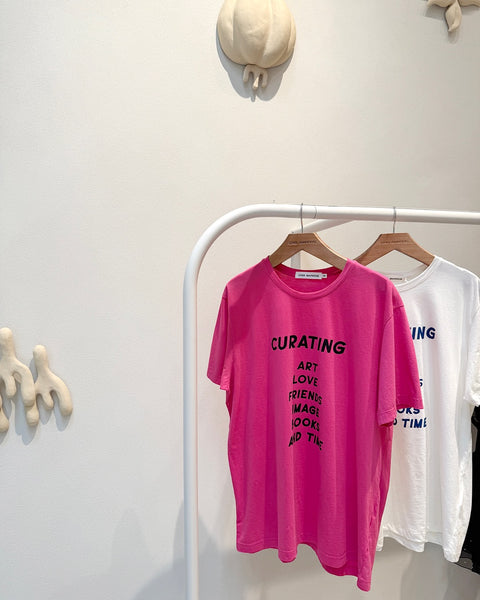 Camisetas da parceira Cris Barros e Act Arte, edição limitada. As camisetas nas cores rosa e off-white estão penduradas em cabides