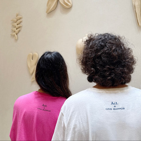 Duas pessoas de costas usando as camisetas da edição limitada da collab da cris barros e act arte, mostrando o detalhe das costas da camiseta